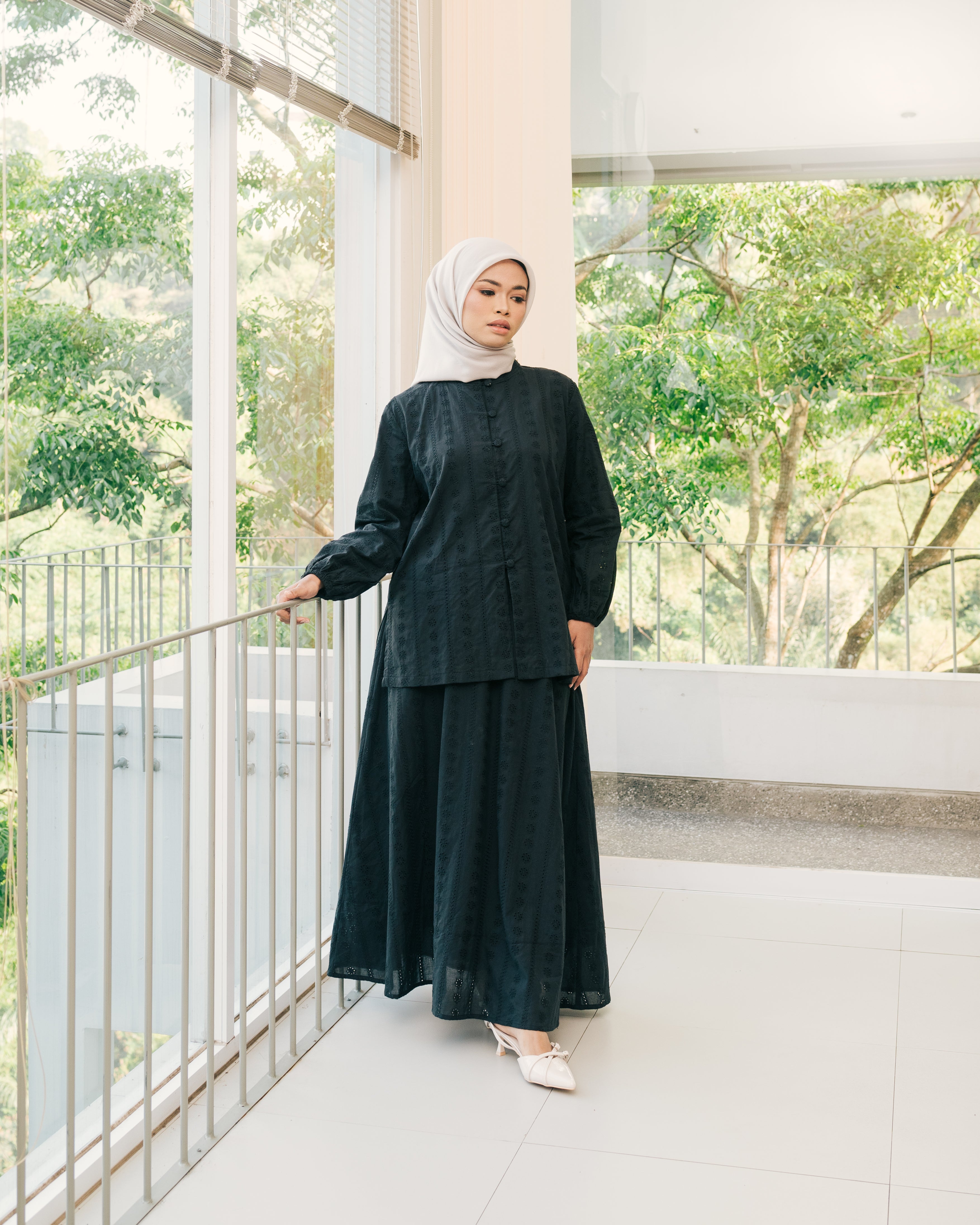 Woman in a black top and skirt from Atala Scarves standing on a balcony with greenery outside