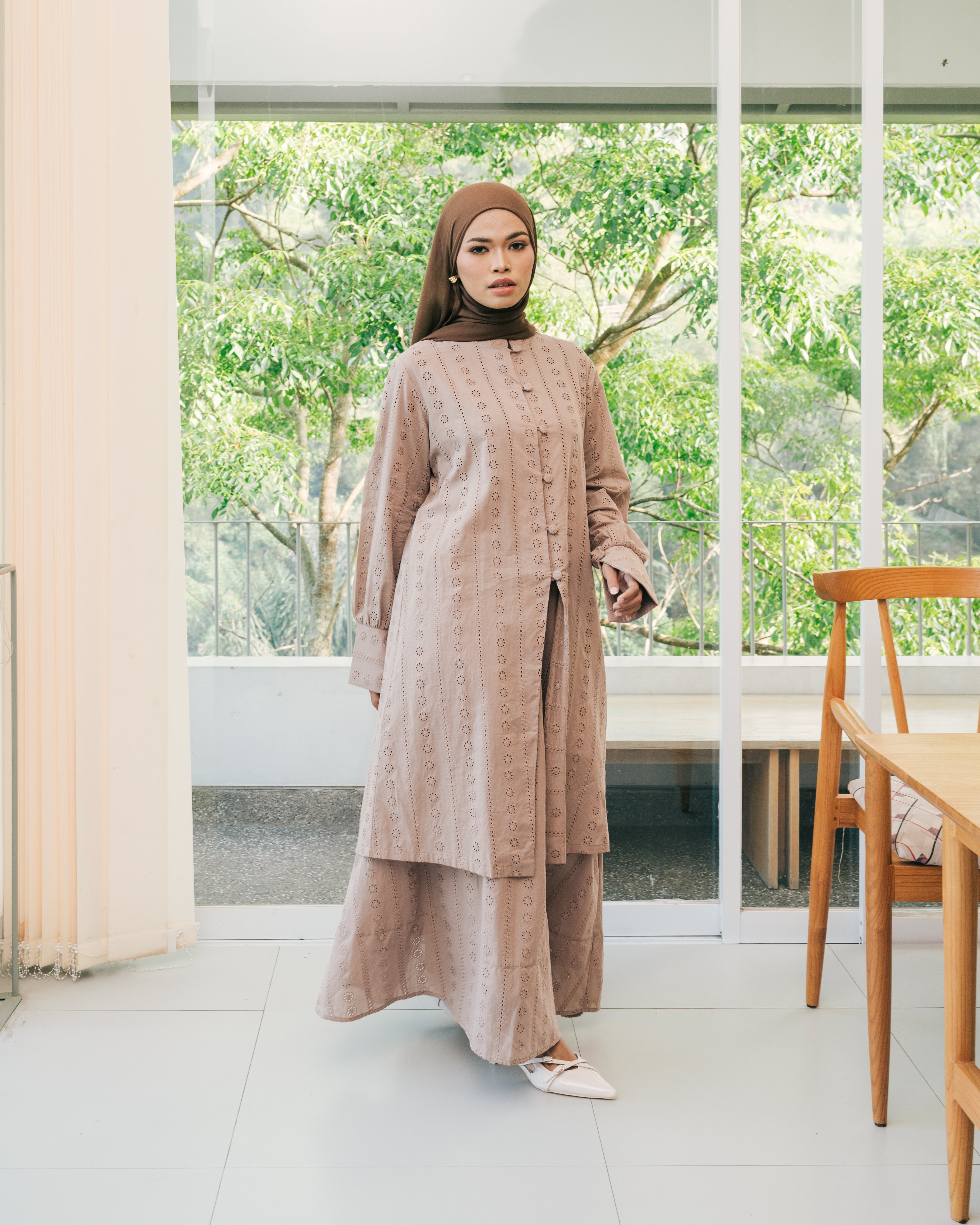 Woman wearing a light brown top and skirt from Atala Scarves standing in a modern room with large windows.