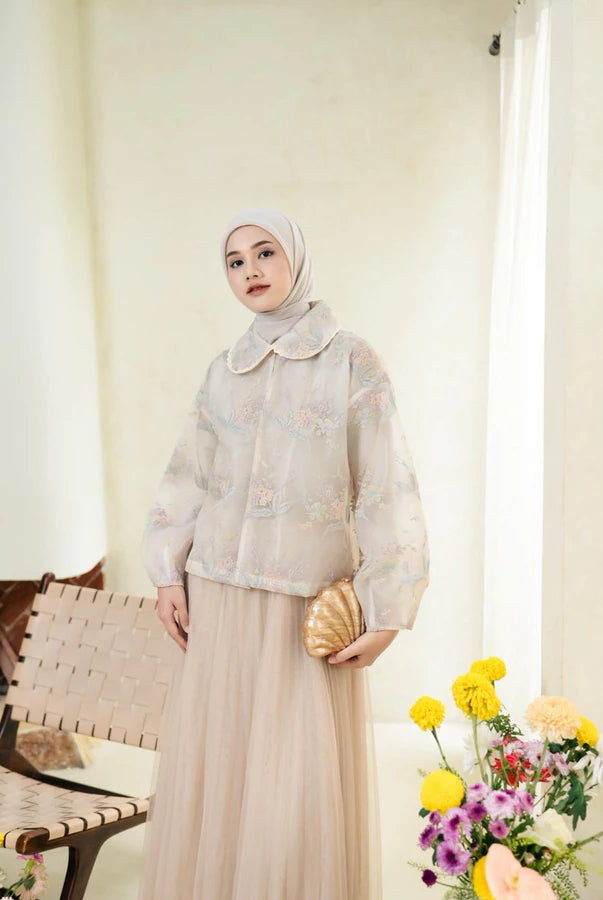 Woman wearing a pink lace outer with floral details by Haidee Orlin with a hijab standing indoors next to a chair and flowers.