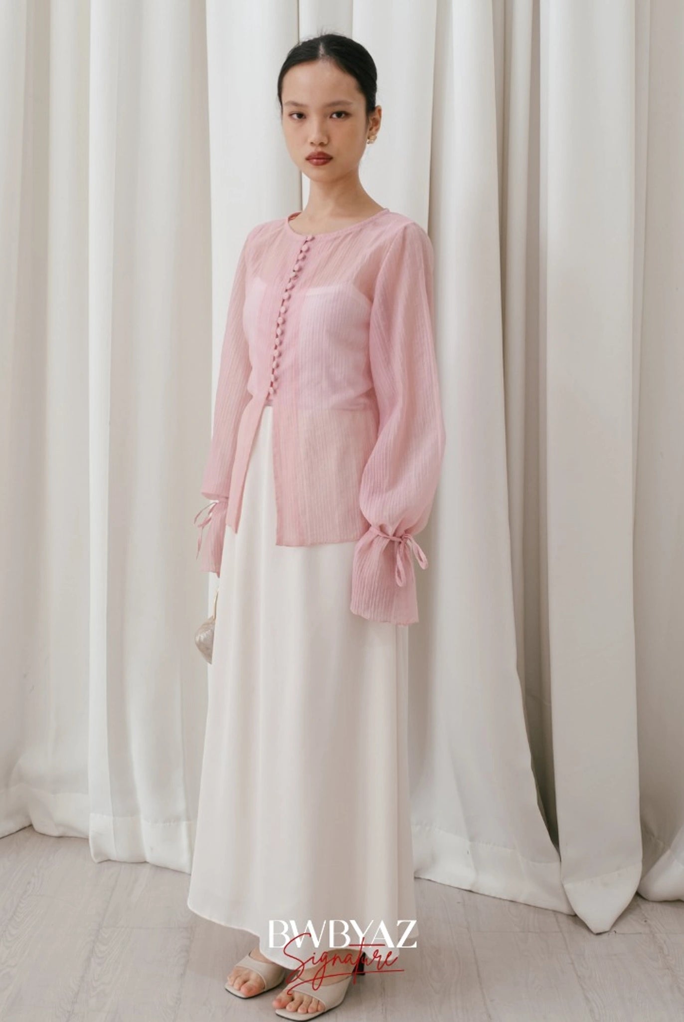 A woman wearing a soft pink, long-sleeved blouse with front buttons and a waist tie from BWBYAZ, paired with a white skirt.