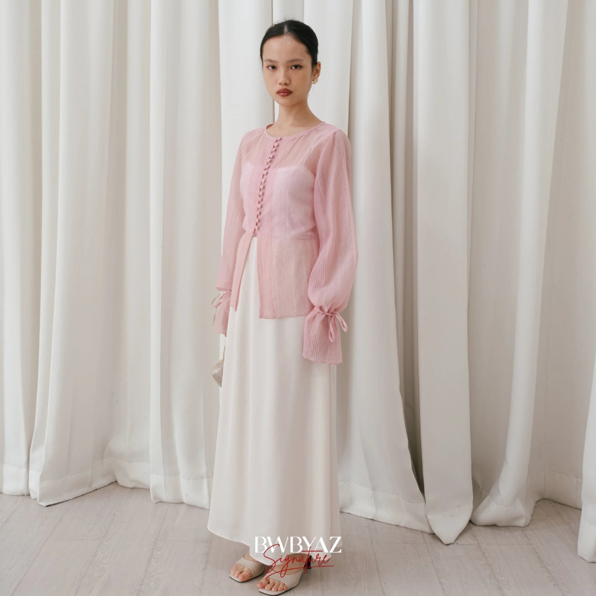 A woman wearing a soft pink, long-sleeved blouse with front buttons and a waist tie from BWBYAZ, paired with a white skirt.