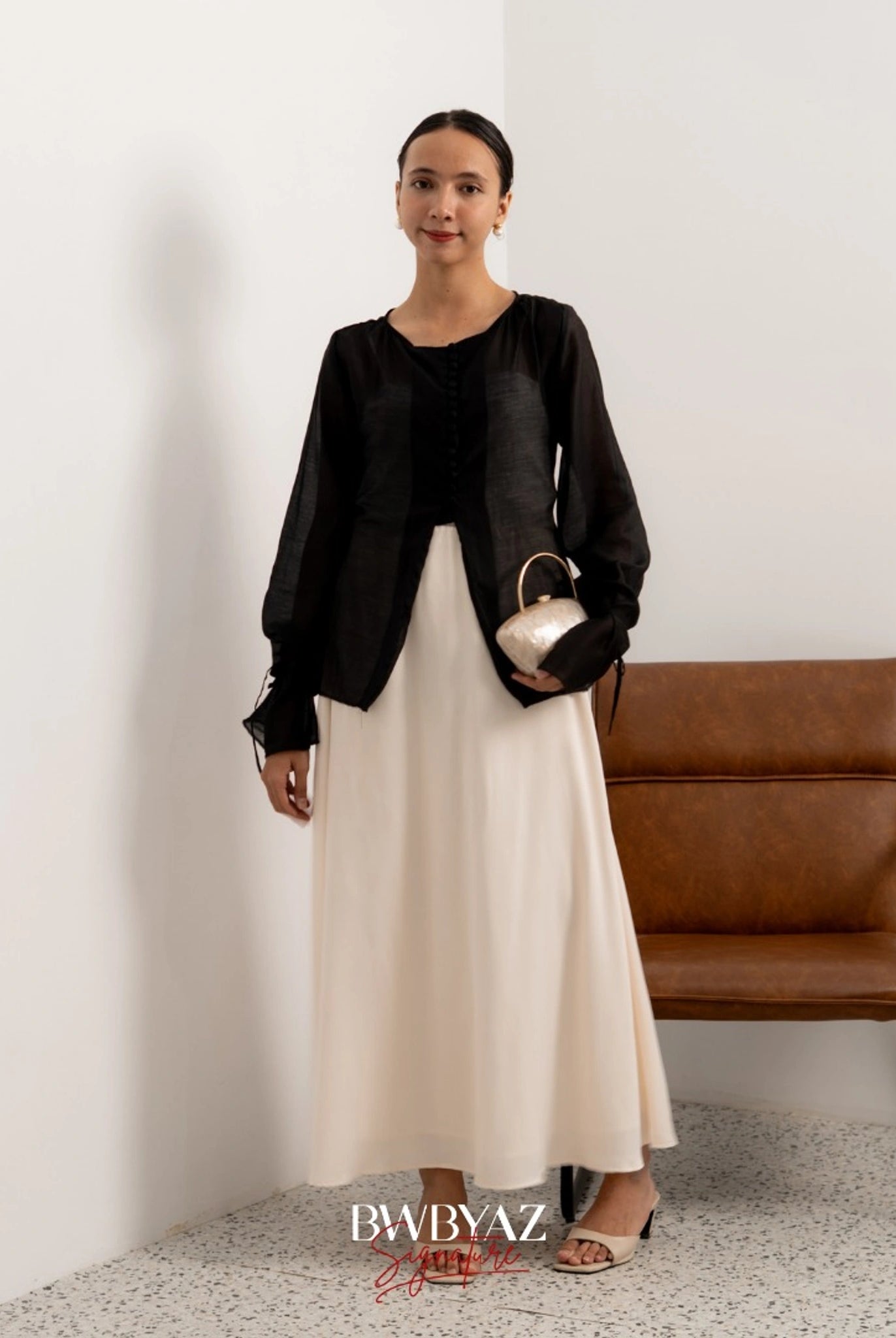 A woman wearing a black button top blouse from BWBYAZ and a sheer beige skirt, standing in a room with a chair and a wall in the background.