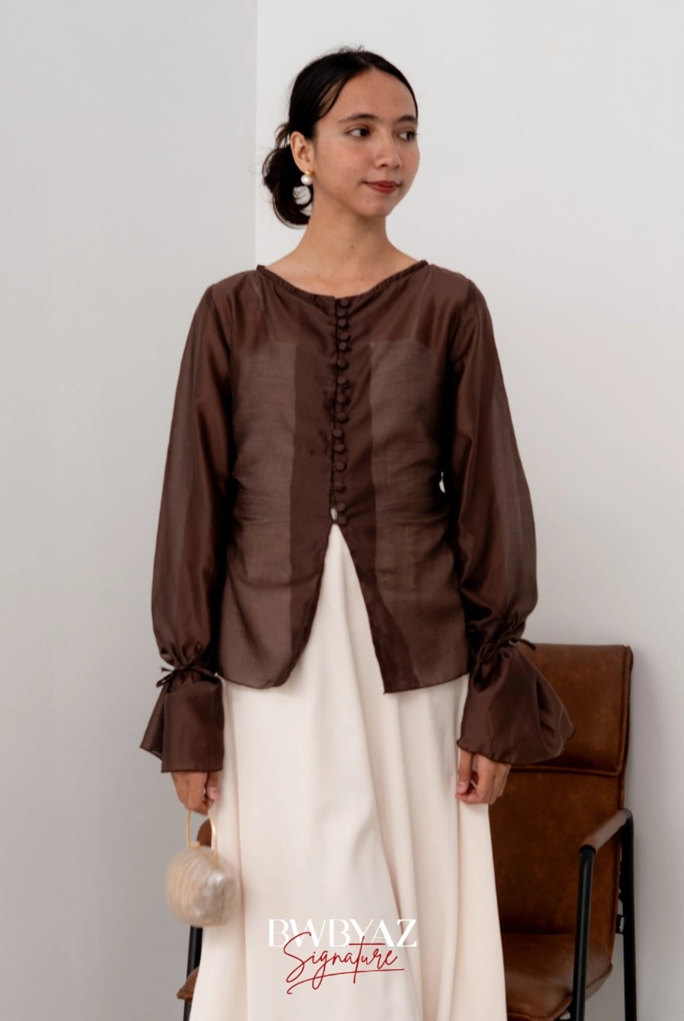 A woman wearing a brown button top blouse from BWBYAZ and a sheer white skirt, standing in a room with a chair and a wall in the background.