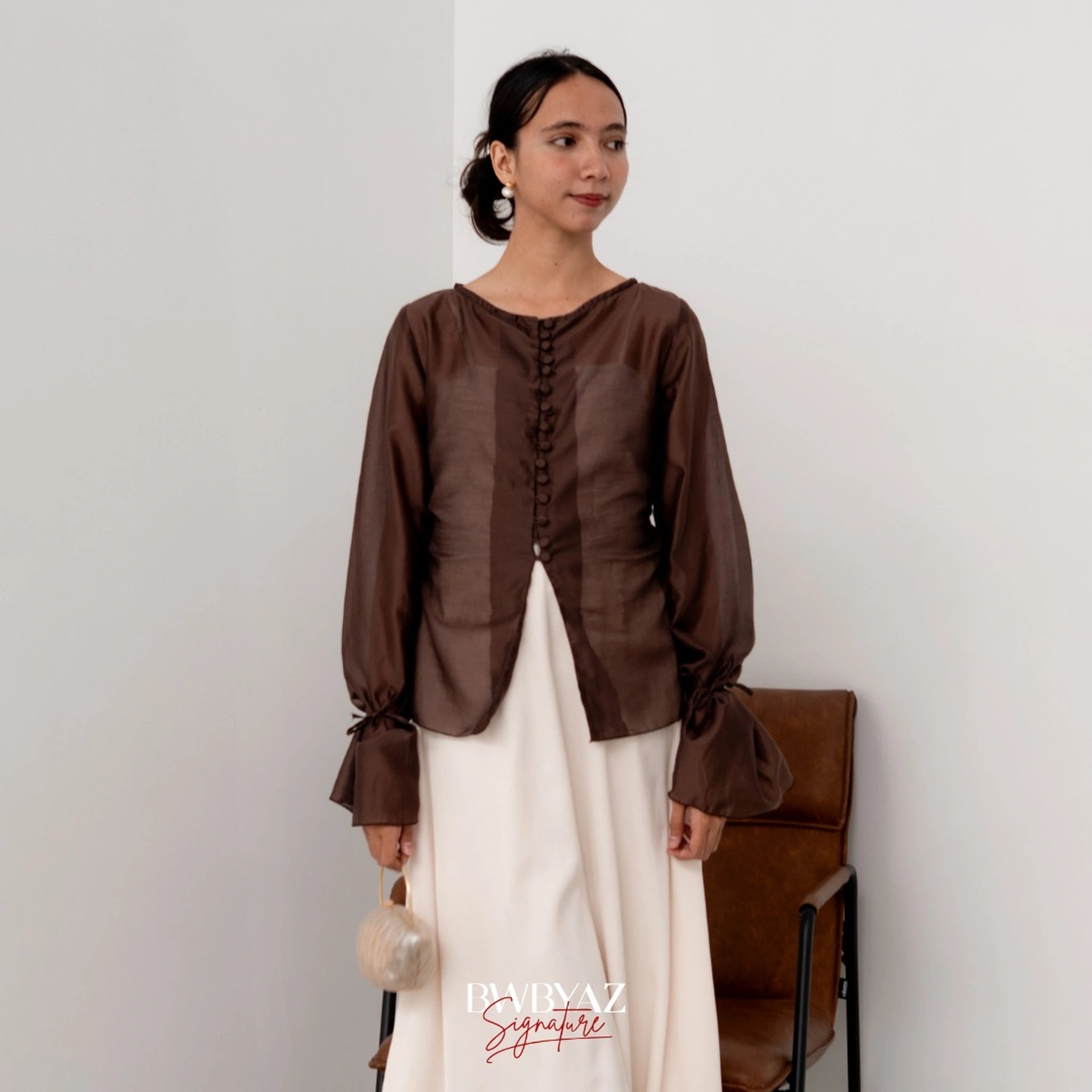 A woman wearing a brown button top blouse from BWBYAZ and a sheer white skirt, standing in a room with a chair and a wall in the background.