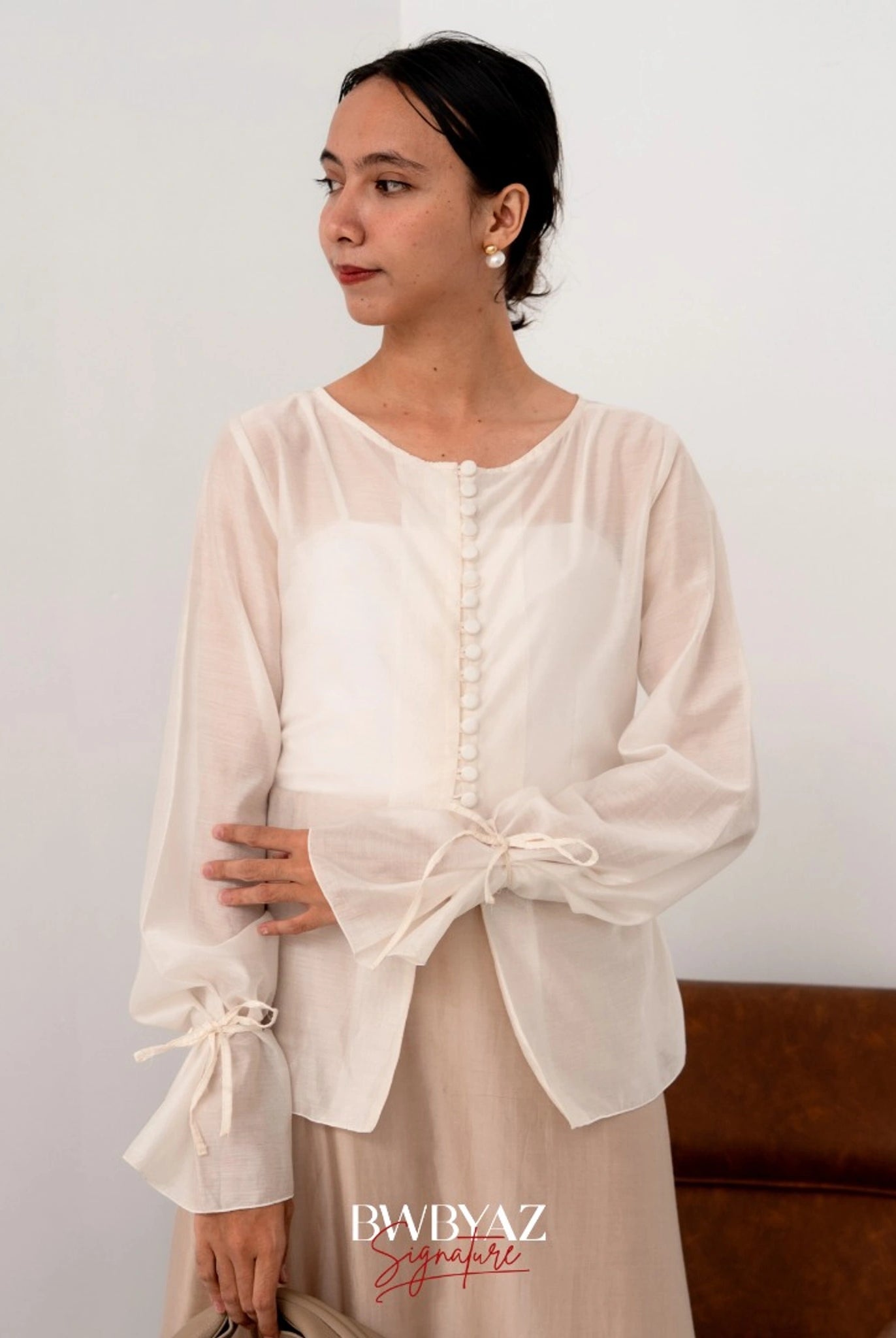 A woman wearing a white button top blouse from BWBYAZ and a sheer beige skirt, standing in a room with a chair and a wall in the background.