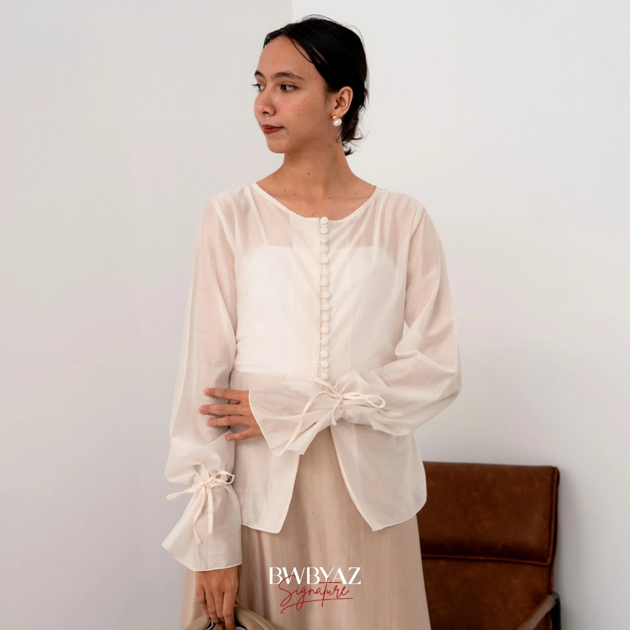 A woman wearing a white button top blouse from BWBYAZ and a sheer beige skirt, standing in a room with a chair and a wall in the background.