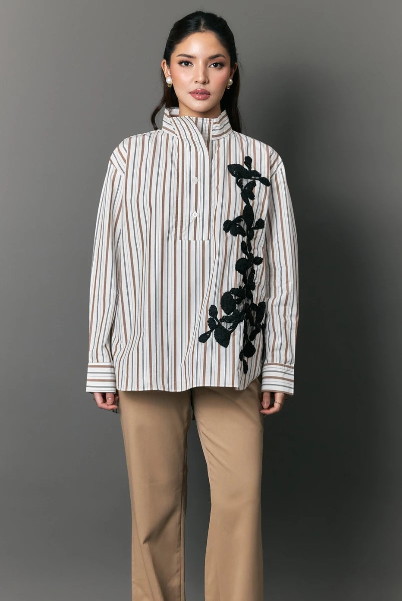Woman wearing a striped shirt with black floral embroidery by Studio Tui and beige pants against a gray background