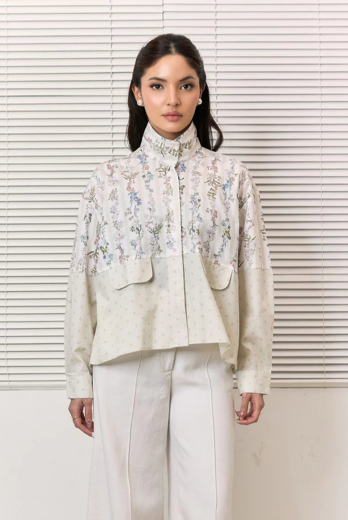 A woman stands in front of white window blinds wearing a high-neck, long-sleeve cropped shirt by Studio Tui with a floral embroidered pattern on the upper portion and solid light beige sleeves, paired with white trousers.