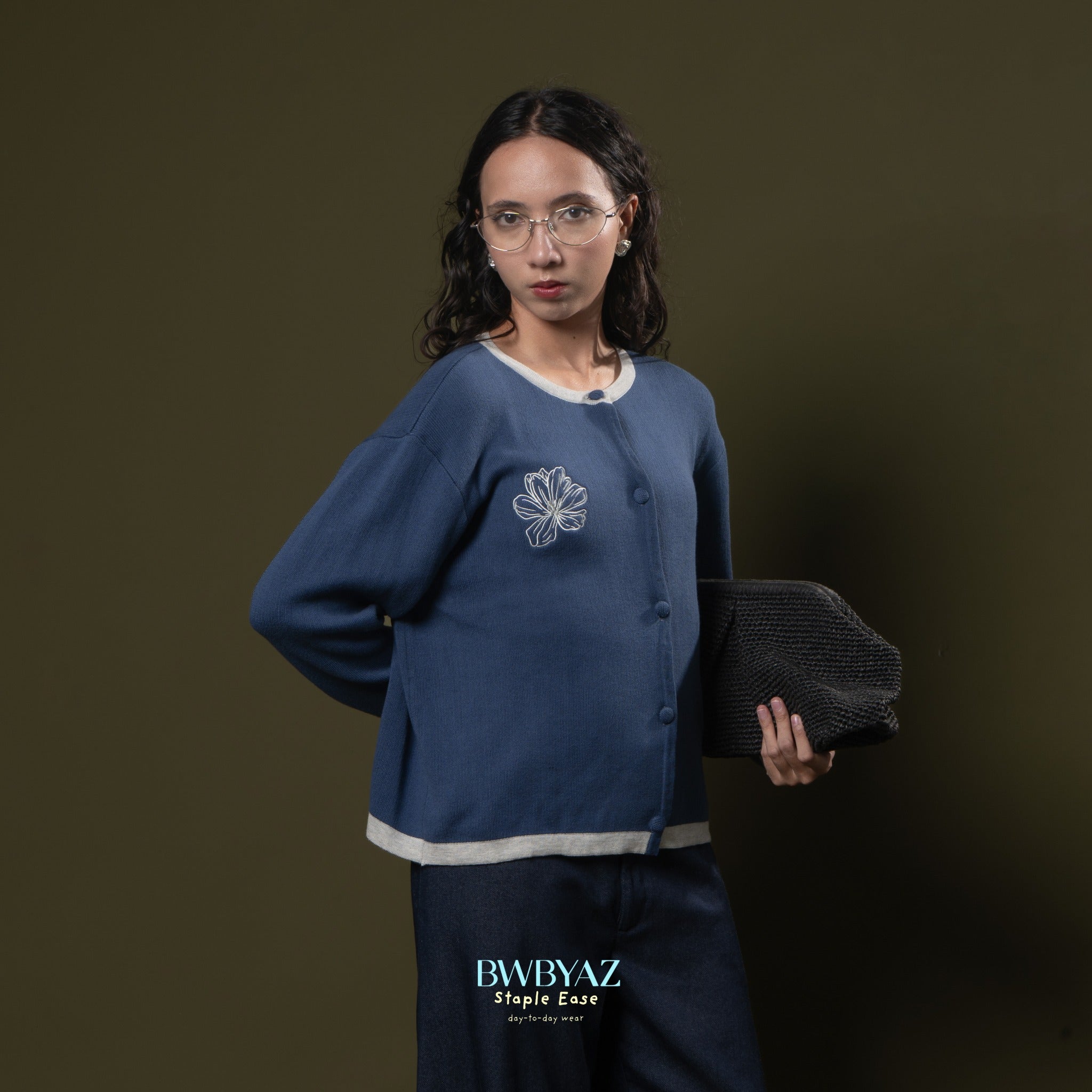 Woman wearing a blue knit cardigan from with a floral design from BWBYAZ, holding a black bag against a dark background.