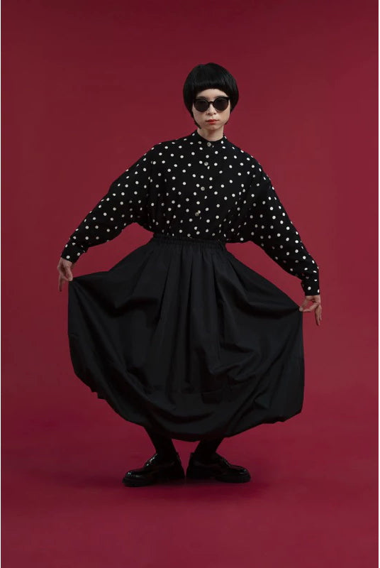 A model wearing a black long-sleeve blouse with white polka dots and a voluminous black skirt by Yestoday, posing against a solid dark red studio background.