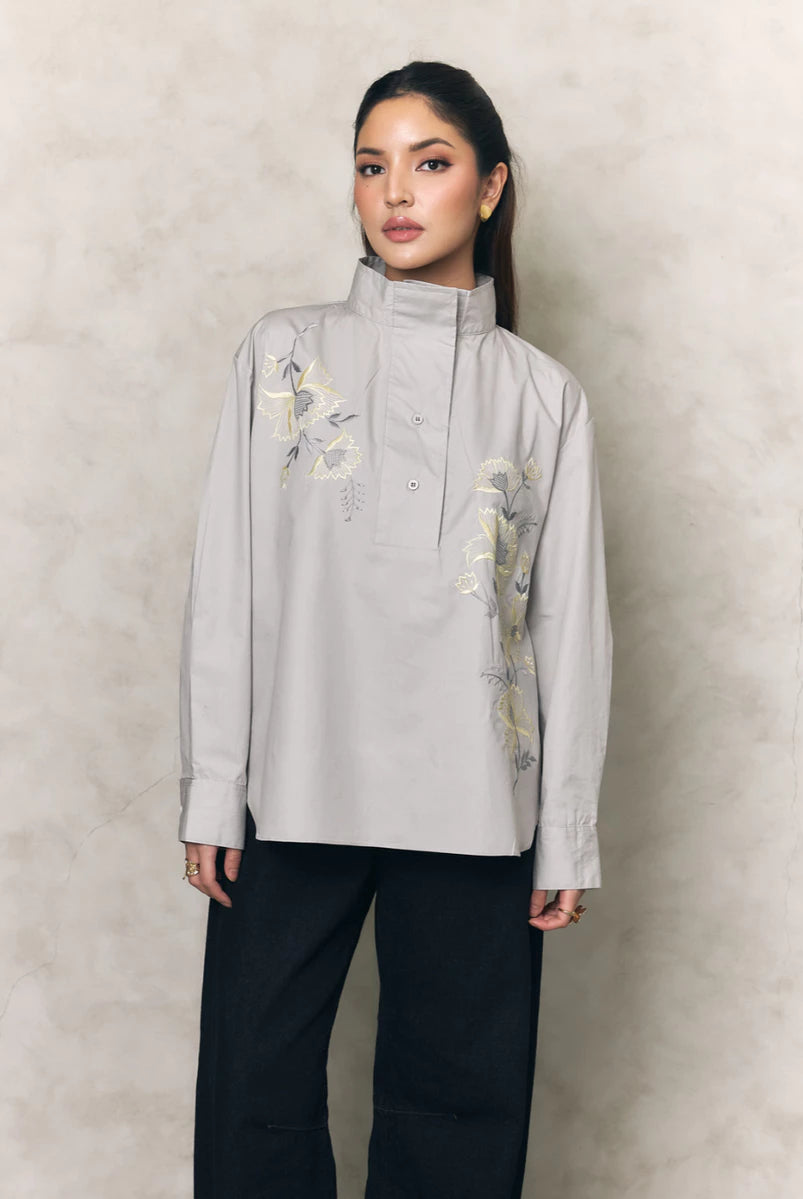 Woman wearing a light gray shirt with floral embroidery by Studio Tui and black pants against a beige background