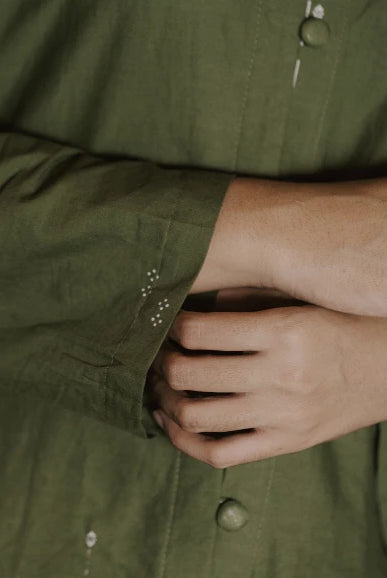 Close-up of a person wearing an olive green button-up tunic, showing the fabric texture, cuff detail with small white embroidered dots, and hands adjusting the shirt.