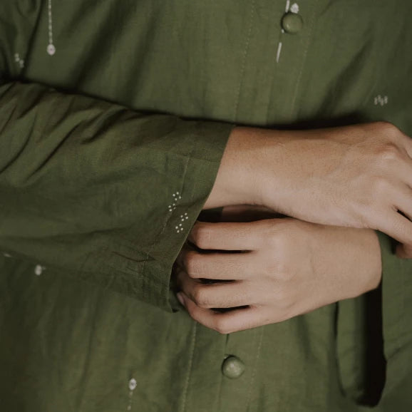 Close-up of a person wearing an olive green button-up tunic, showing the fabric texture, cuff detail with small white embroidered dots, and hands adjusting the shirt.