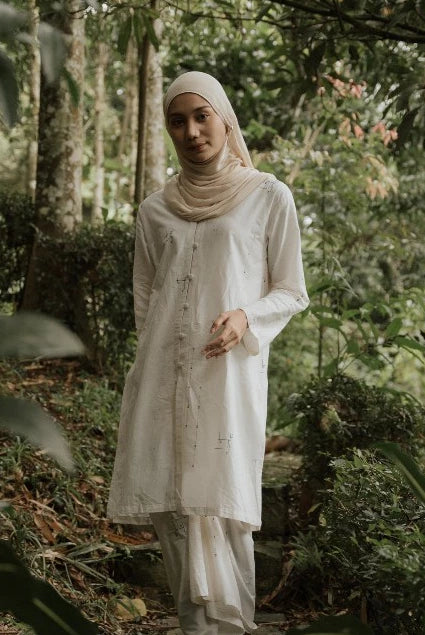 A woman wearing a white long-sleeve tunic and matching pants with a beige hijab stands in a lush green forest.