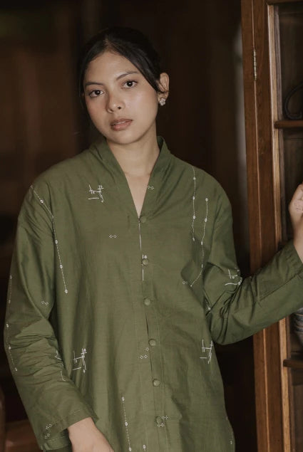 A woman wearing an olive green long-sleeve button-up tunic with white embroidered details, standing in a rustic indoor setting next to a wooden cabinet.