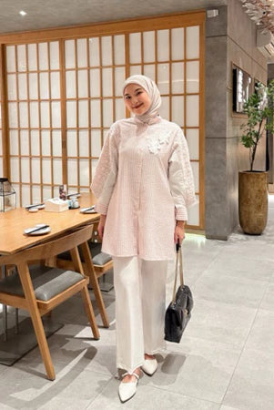 A woman wearing a pink tulip shirt with long sleeves by Casa Elana, paired with white trousers, standing in a room with wooden dividers and a table with chairs in the background.