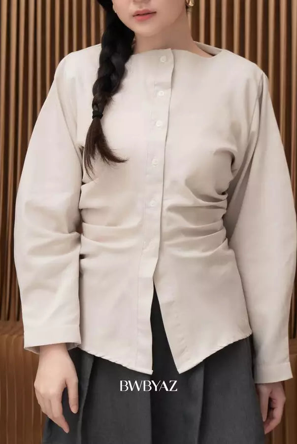 A woman wearing a light beige long-sleeve button-up shirt by BWBYAZ with gathered side details and a boat neckline, paired with dark gray pants, set against a brown slatted wooden background.
