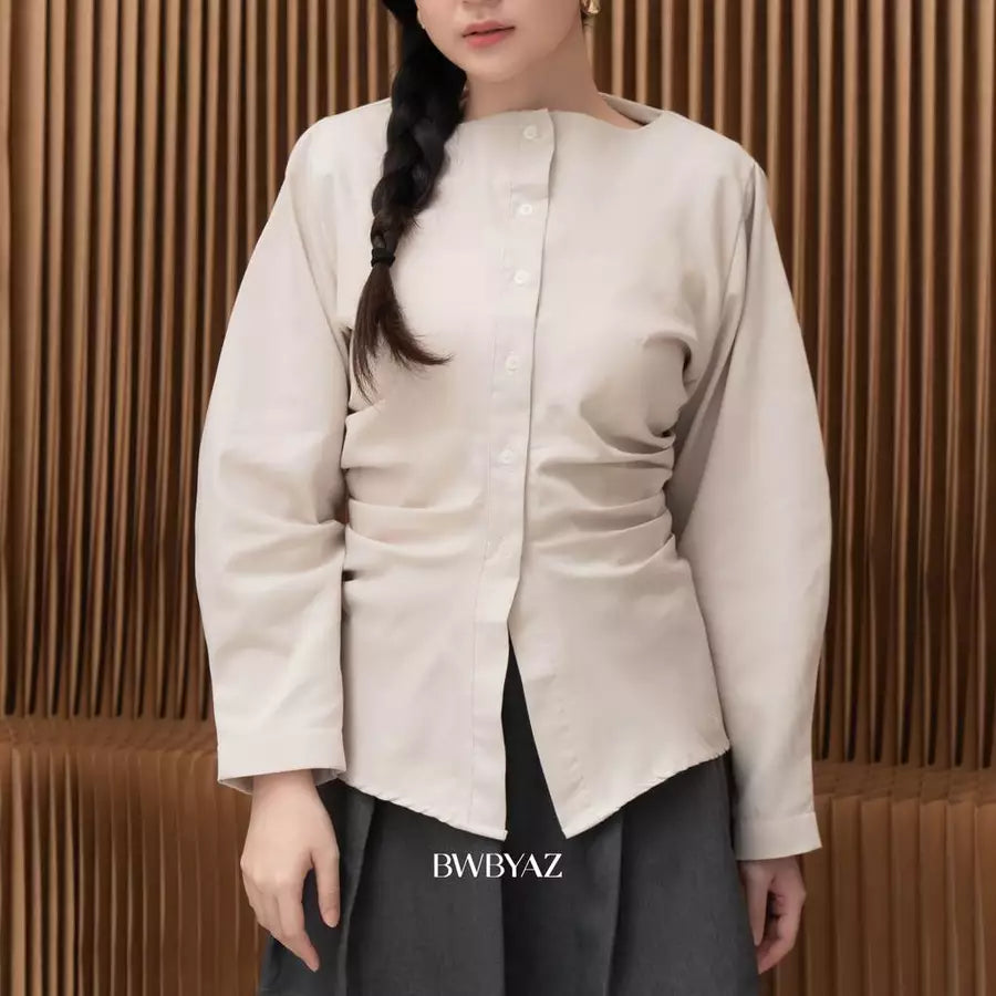 A woman wearing a light beige long-sleeve button-up shirt by BWBYAZ with gathered side details and a boat neckline, paired with dark gray pants, set against a brown slatted wooden background.