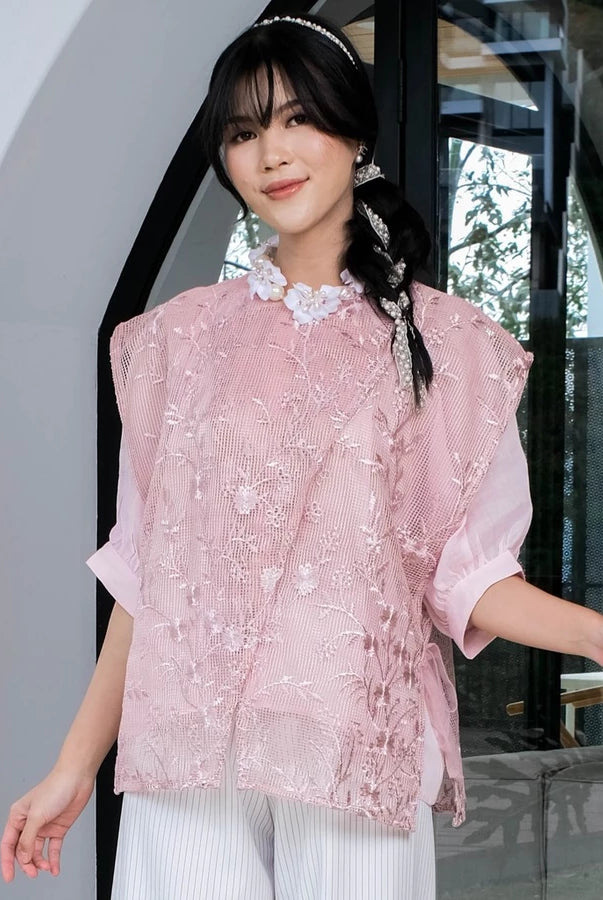 Woman wearing a pink embroidered lace outer by Holy Peony and white pants standing indoors.