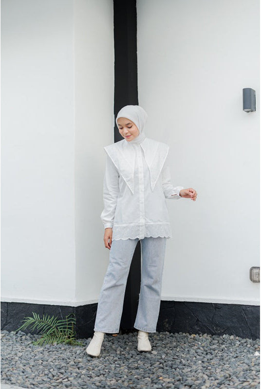 Woman wearing a white layered long-sleeve shirt by Casa Elana, light blue jeans, and a white hijab standing against a minimalist white wall.