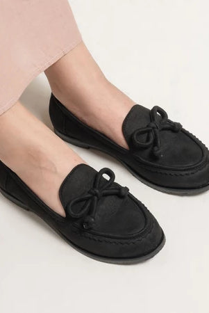 Black loafers flat shoes with bow details from Dianable worn by a person on a light background