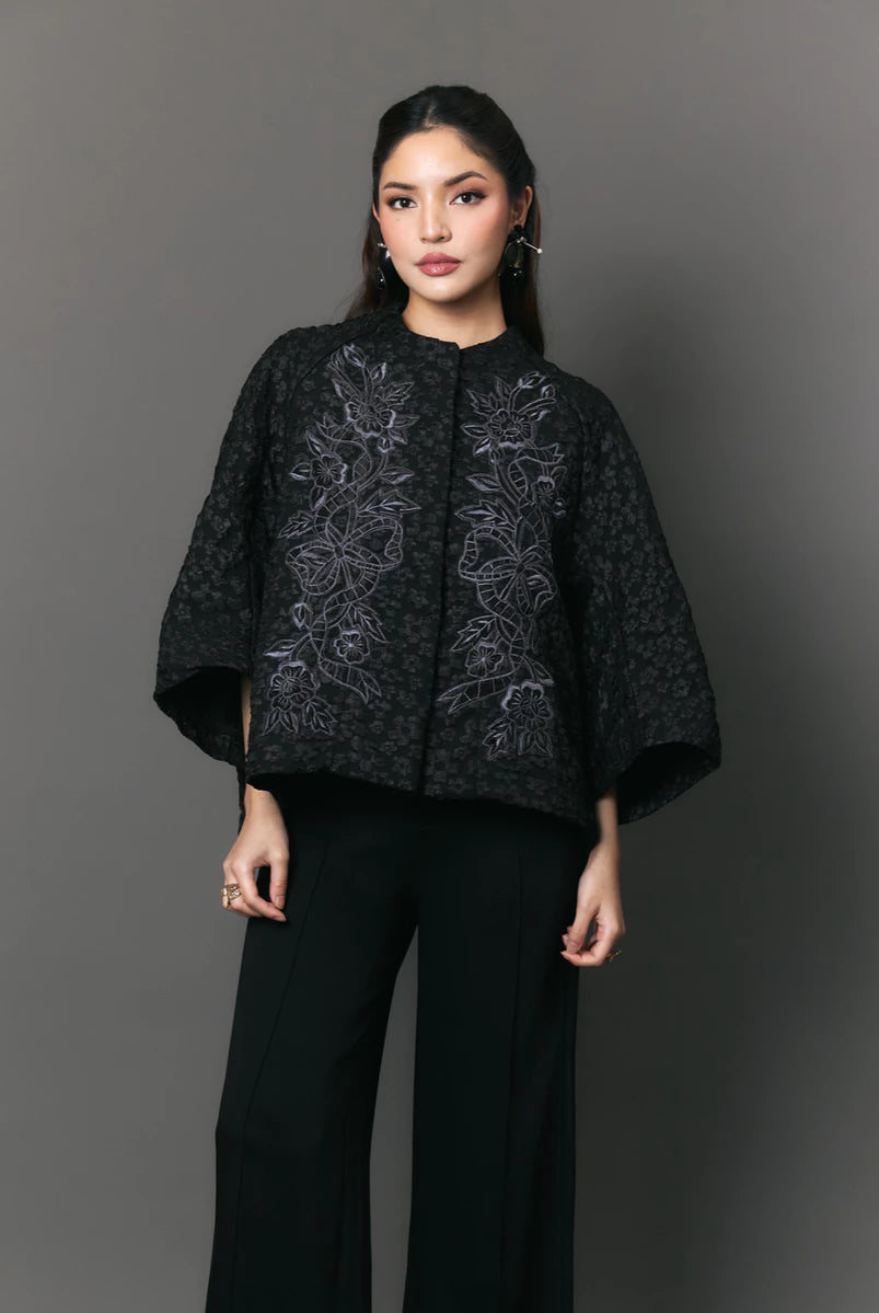 Woman wearing a black patterned outer with floral embriodery by Studio Tui against a gray background