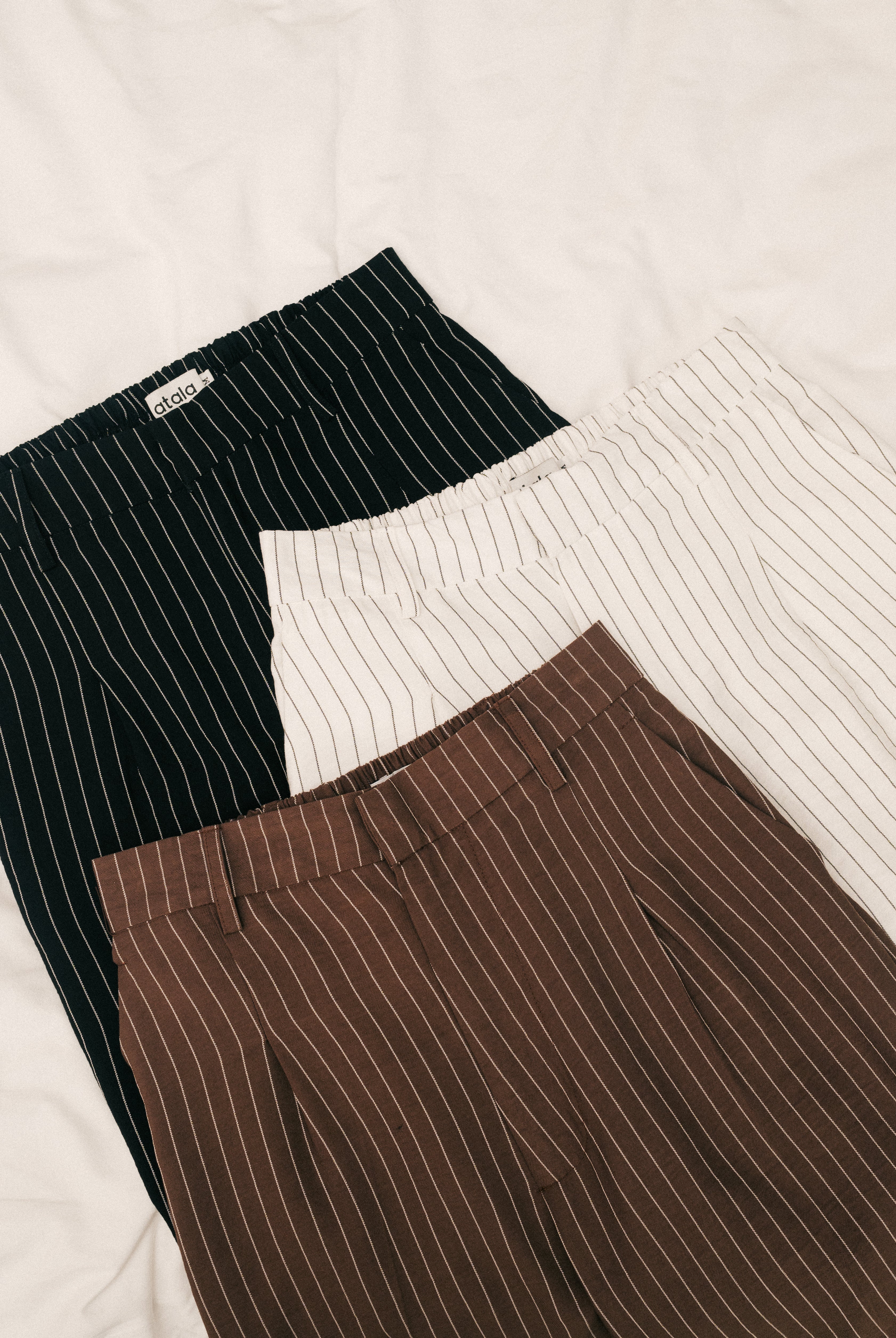 Three pairs of pinstripe pants in black, white, and brown from Atala Scarves on a light background.