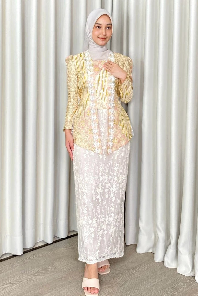 A woman wearing a traditional white skirt by Kartinis Label with floral patterns, standing in front of a curtain.