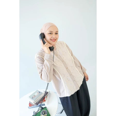 Woman wearing a beige hijab and shirt by Abame, holding a black phone receiver against a white background.