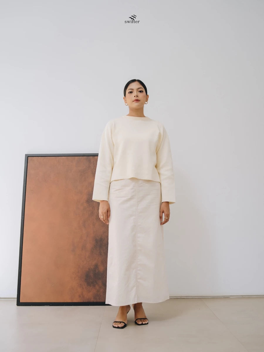 Woman wearing a broken white-colored knit sweater by Swifter standing against a white wall with a framed piece of art.