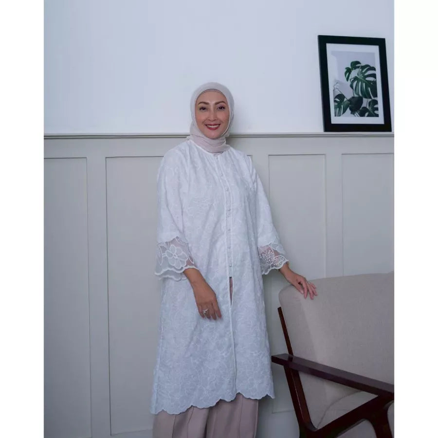 Woman wearing a white lace tunic by Abame standing in a room with a white wall and a framed picture.
Seorang wanita mengenakan tunik renda putih dari Abame berdiri di dalam ruangan dengan dinding putih dan sebuah foto dalam bingkai.