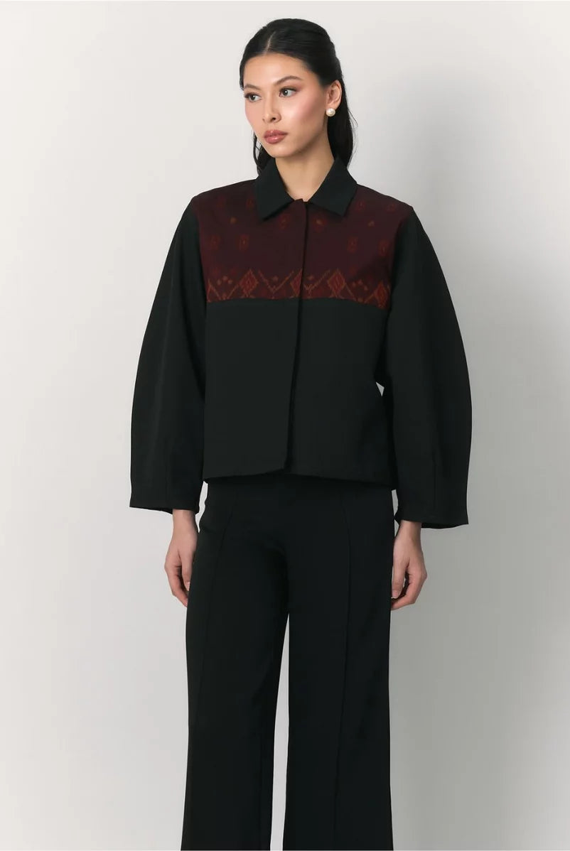 A model wearing a black cropped jacket by Studio Tui with a dark red patterned yoke and black wide-leg trousers, photographed against a light gray studio background.