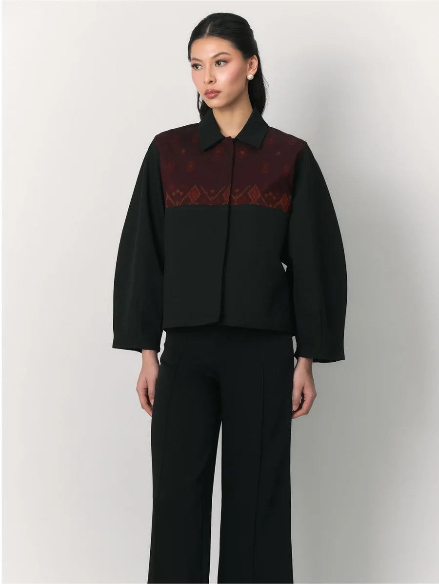 A model wearing a black cropped jacket by Studio Tui with a dark red patterned yoke and black wide-leg trousers, photographed against a light gray studio background.