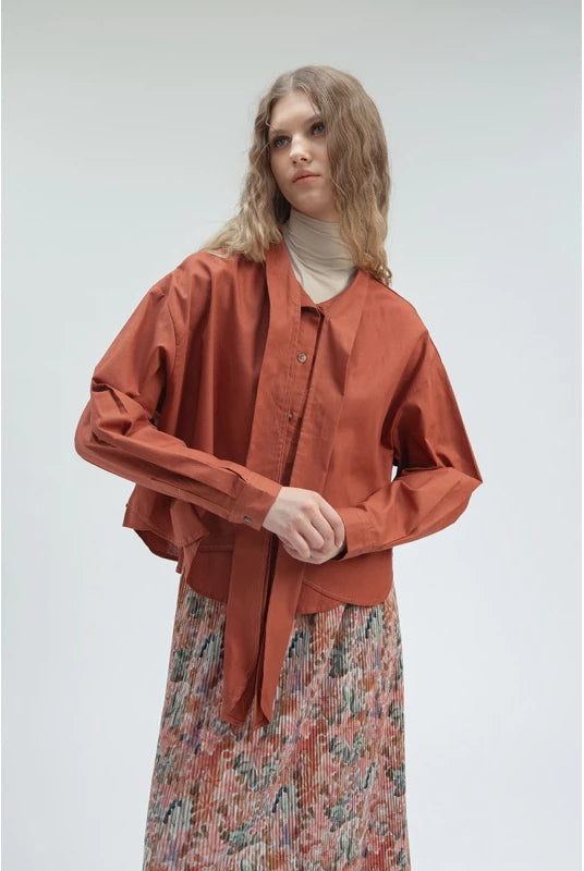 Woman wearing a terracotta women’s tie blouse by Yestoday with a loose silhouette and front button details, paired with a cream turtleneck inner and a floral pleated skirt for a modern feminine look.