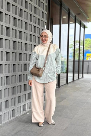 Woman in hijab wearing a green stripe shirt by Abame standing against a modern building with glass windows.