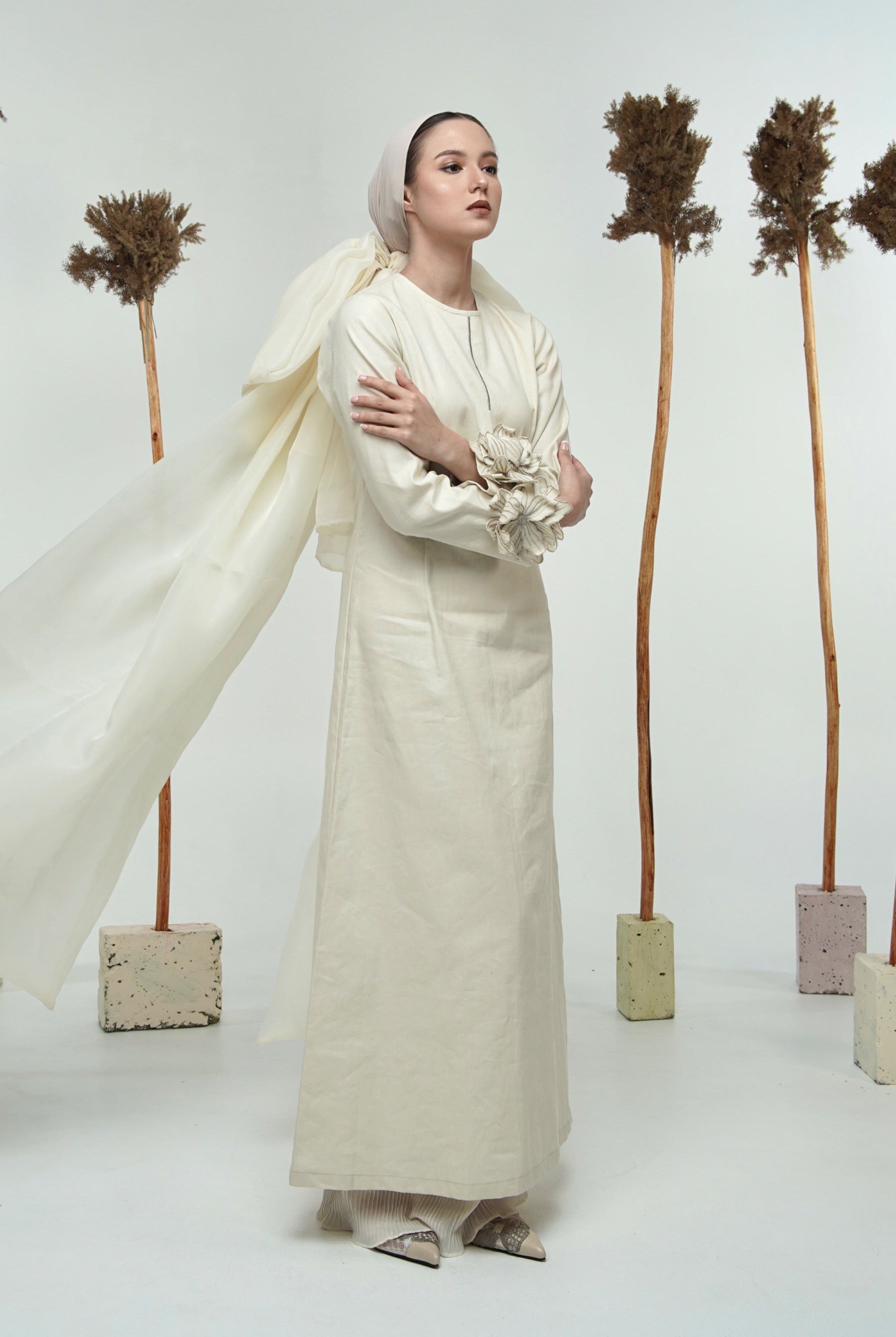 A model wearing a long white linen dress with long sleeves by Glashka, standing against a backdrop with decorative trees.
