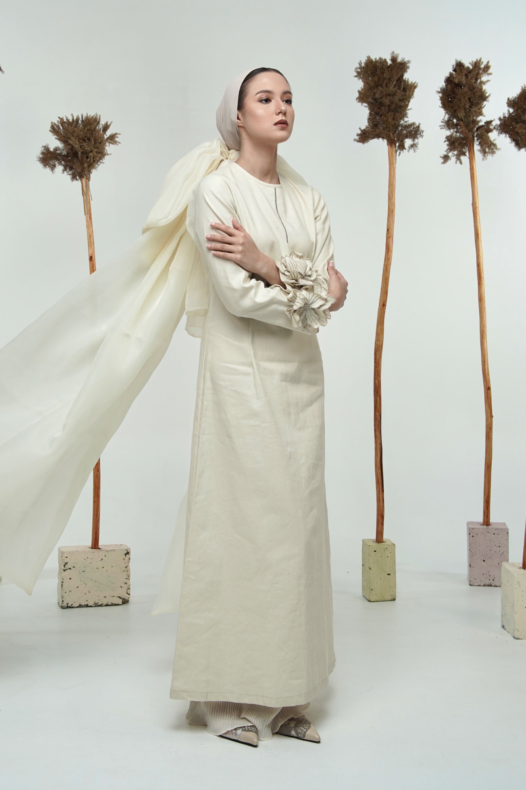 A model wearing a long white linen dress with long sleeves by Glashka, standing against a backdrop with decorative trees.