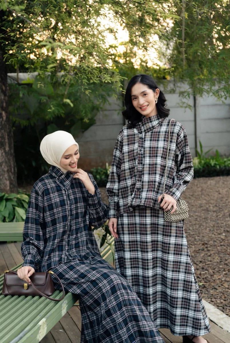 Two women posing outdoors in matching long-sleeve plaid outfits by Haidee Orlin. One is seated on a green bench wearing a white hijab and holding a brown handbag, while the other stands beside her in a dark plaid long-sleeve top and skirt set.