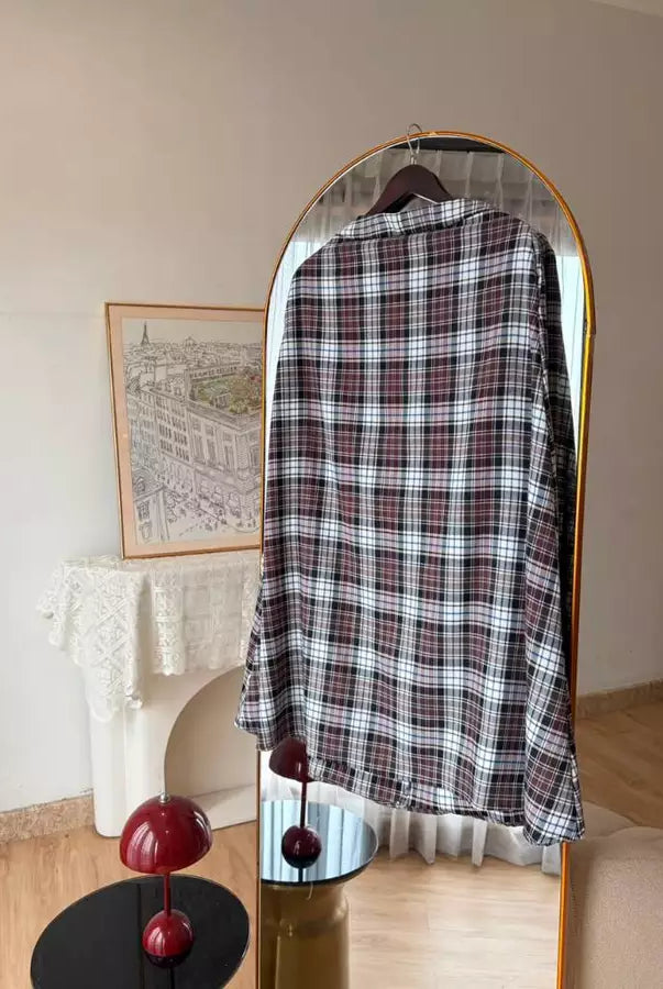 A long-sleeve plaid shirt in black, white, and red by Haidee Orlin is displayed on a hanger inside a gold-framed arched mirror, set against a warm beige wall with a white fireplace mantel in the background.