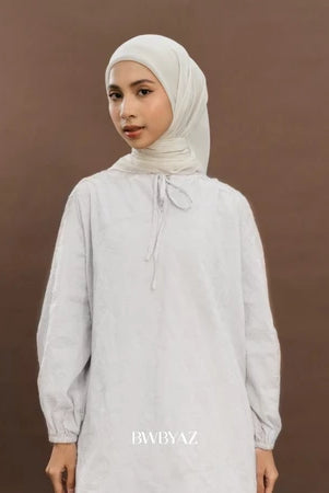 A woman wearing a white long tunic with floral pattern, featuring tie details at the neckline and bishop sleeves with elastic cuffs from BWBYAZ.