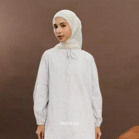 A woman wearing a white long tunic with floral pattern, featuring tie details at the neckline and bishop sleeves with elastic cuffs from BWBYAZ.