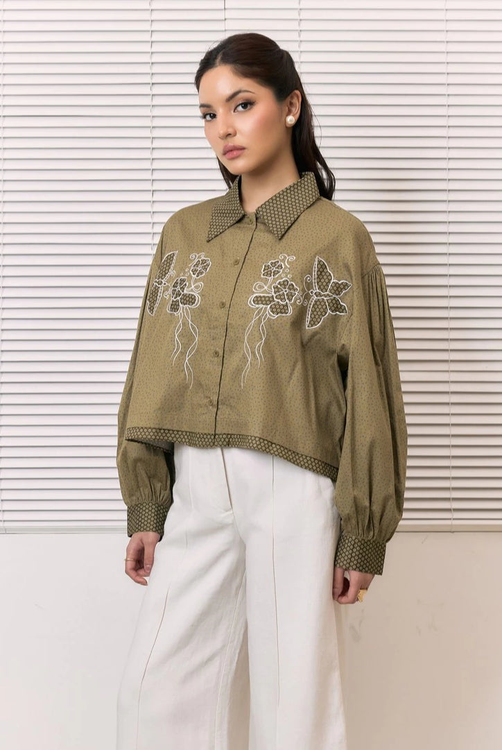 A woman models a cropped, long-sleeve button-up shirt in olive green by Studio Tui with white floral embroidery on the front, paired with white wide-leg pants, set against a background of white window blinds.