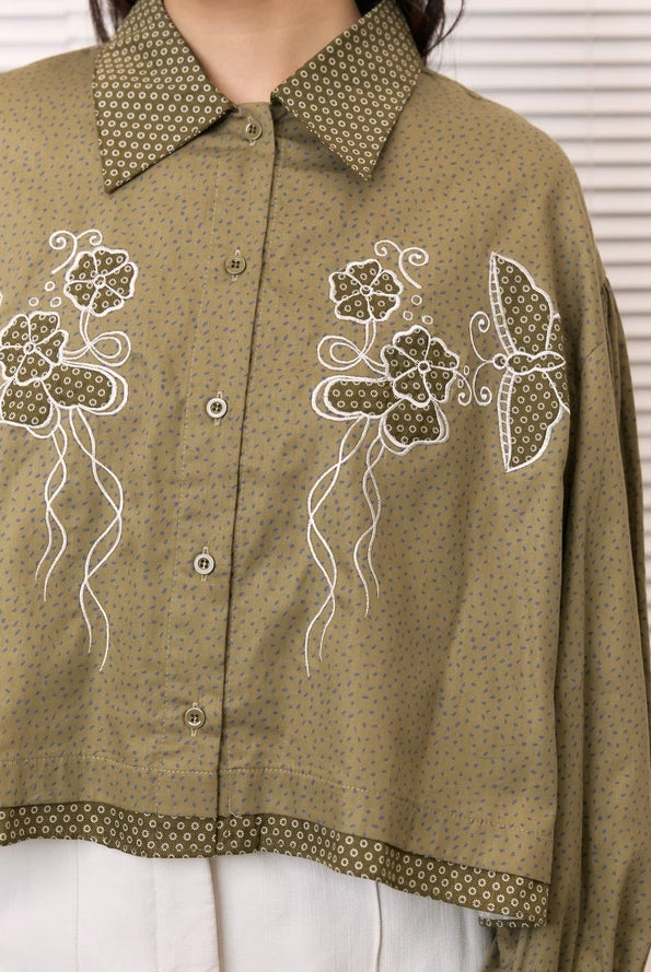 An olive green cropped button-up shirt by Studio Tui with a fine blue dot pattern, featuring white embroidered floral and butterfly designs on the front, worn by a model.