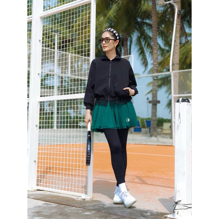 Person on a tennis court wearing a black track jacket by Haidee Orlin, green skirt, and white shoes.