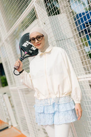 Person wearing an ivory-colored track jacket by Haidee Orlin and hijab, standing on a tennis court.