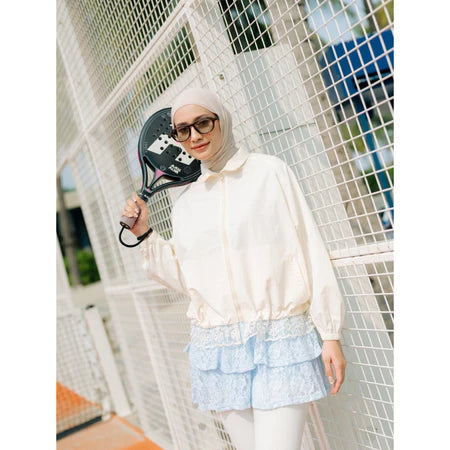 Person wearing an ivory-colored track jacket by Haidee Orlin and hijab, standing on a tennis court.