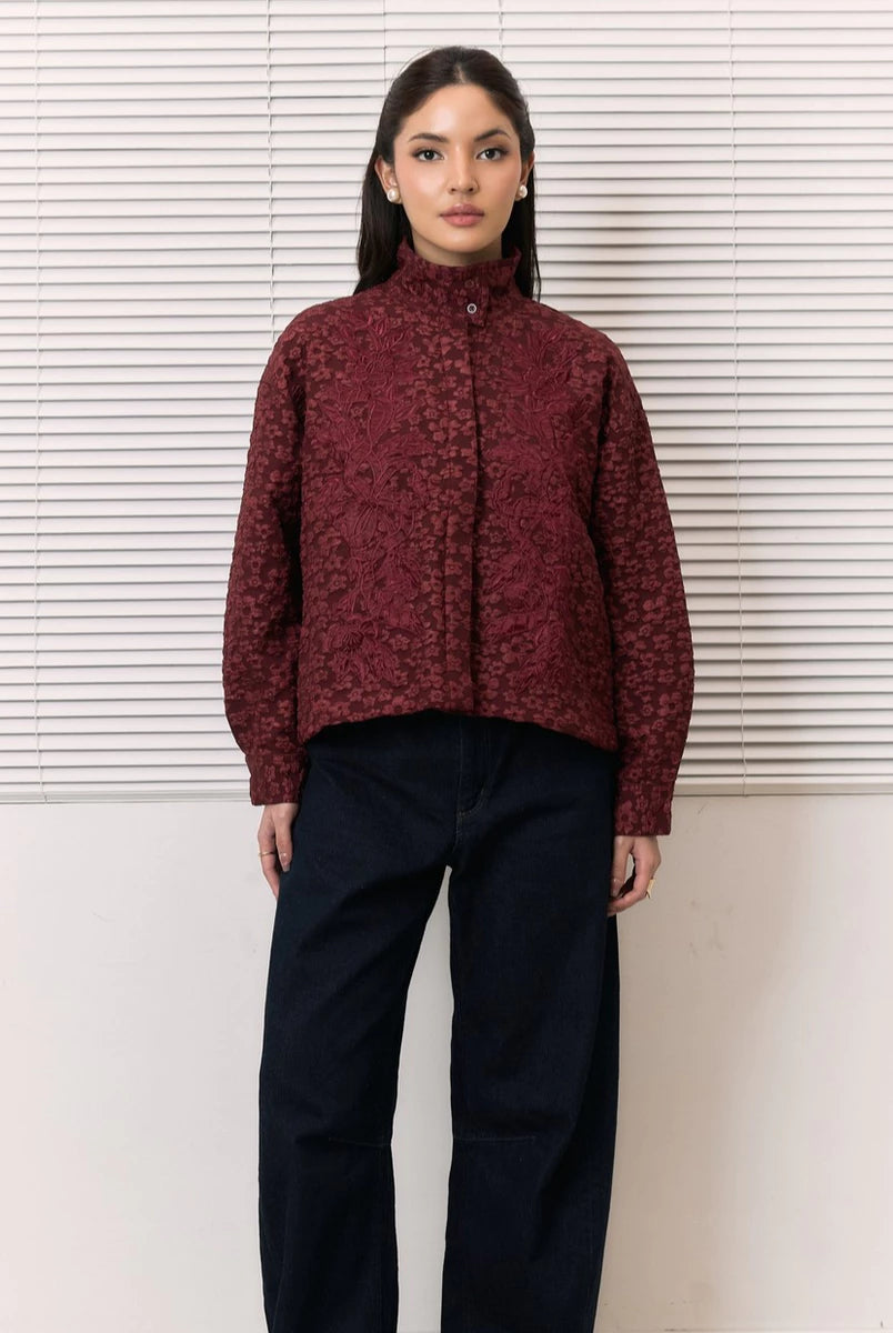 A woman stands in front of white horizontal blinds wearing a cropped, long-sleeve high-neck shirt by Studio Tui in a dark burgundy floral jacquard pattern, paired with dark navy wide-leg pants.