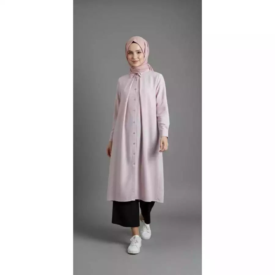 Woman wearing a light pink tunic and black pants on a gray background.
Seorang wanita mengenakan tunik berwarna pink muda dan celana hitam di latar belakang abu-abu.