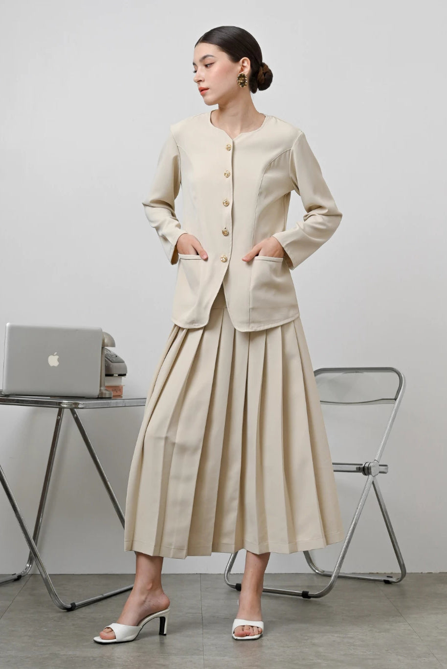 A model wearing a beige Zora skirt by Kasa Heritage with pleats, paired with a matching brown top. The skirt reaches mid-calf and has a fitted waist with a zipper at the back.