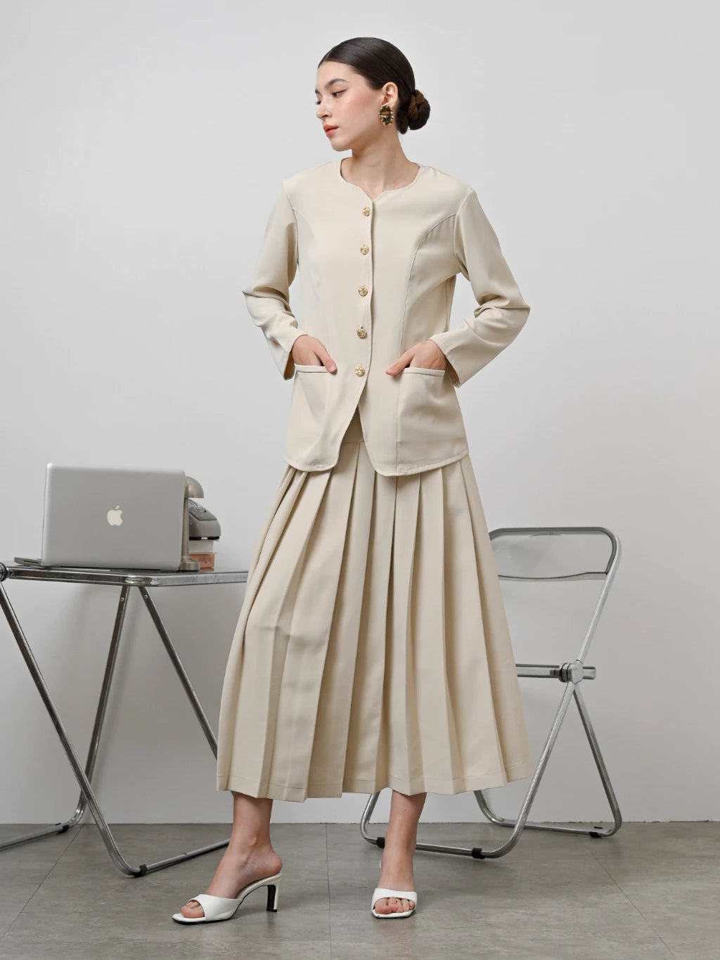 A model wearing a beige Zora skirt by Kasa Heritage with pleats, paired with a matching brown top. The skirt reaches mid-calf and has a fitted waist with a zipper at the back.